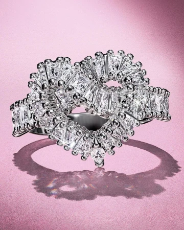 Close-up of a heart-shaped diamond ring featuring baguette-cut diamonds and bead details, set against a soft pink background.