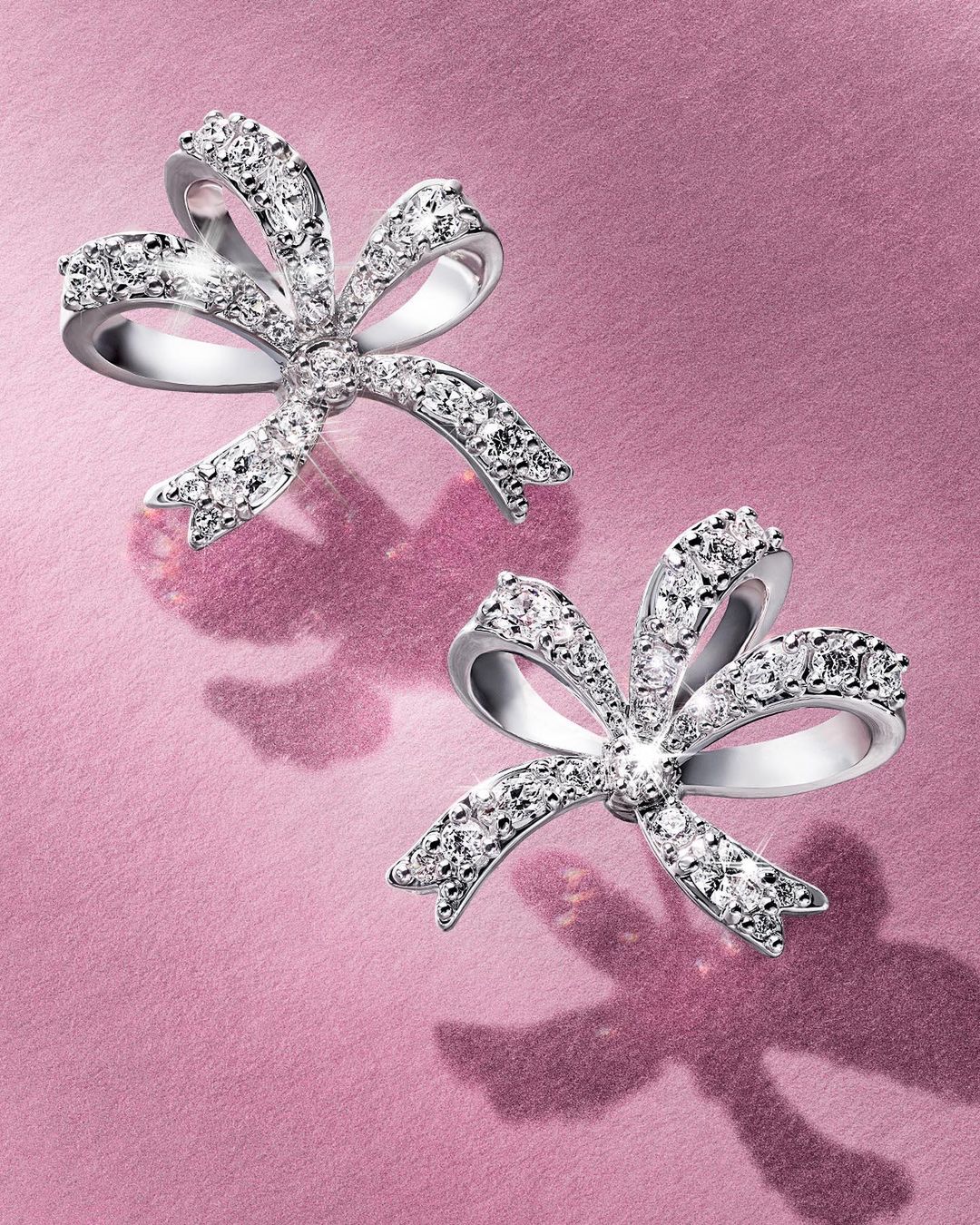 Two diamond bow earrings displayed on a shimmering textured pink background with soft shadows