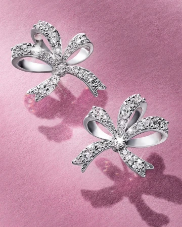 Two diamond bow earrings displayed on a shimmering textured pink background with soft shadows