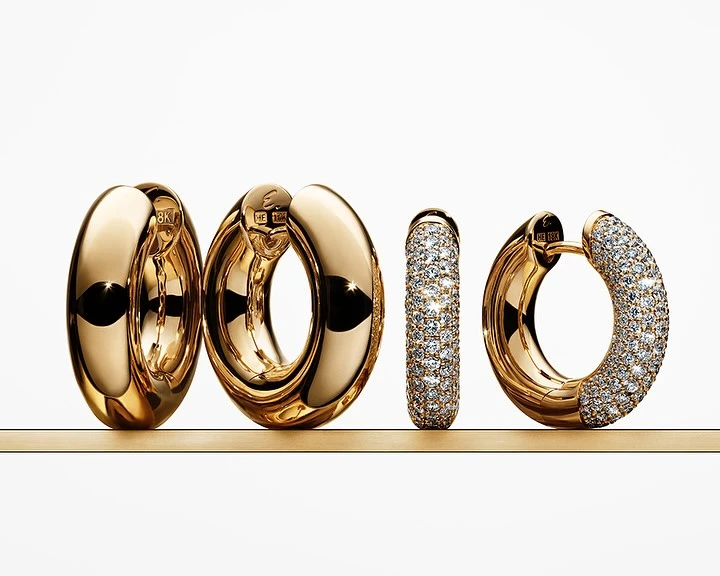 Close-up studio shot of gold hoop earrings, some partially encrusted with diamonds, displayed on a gold bar against a white background.