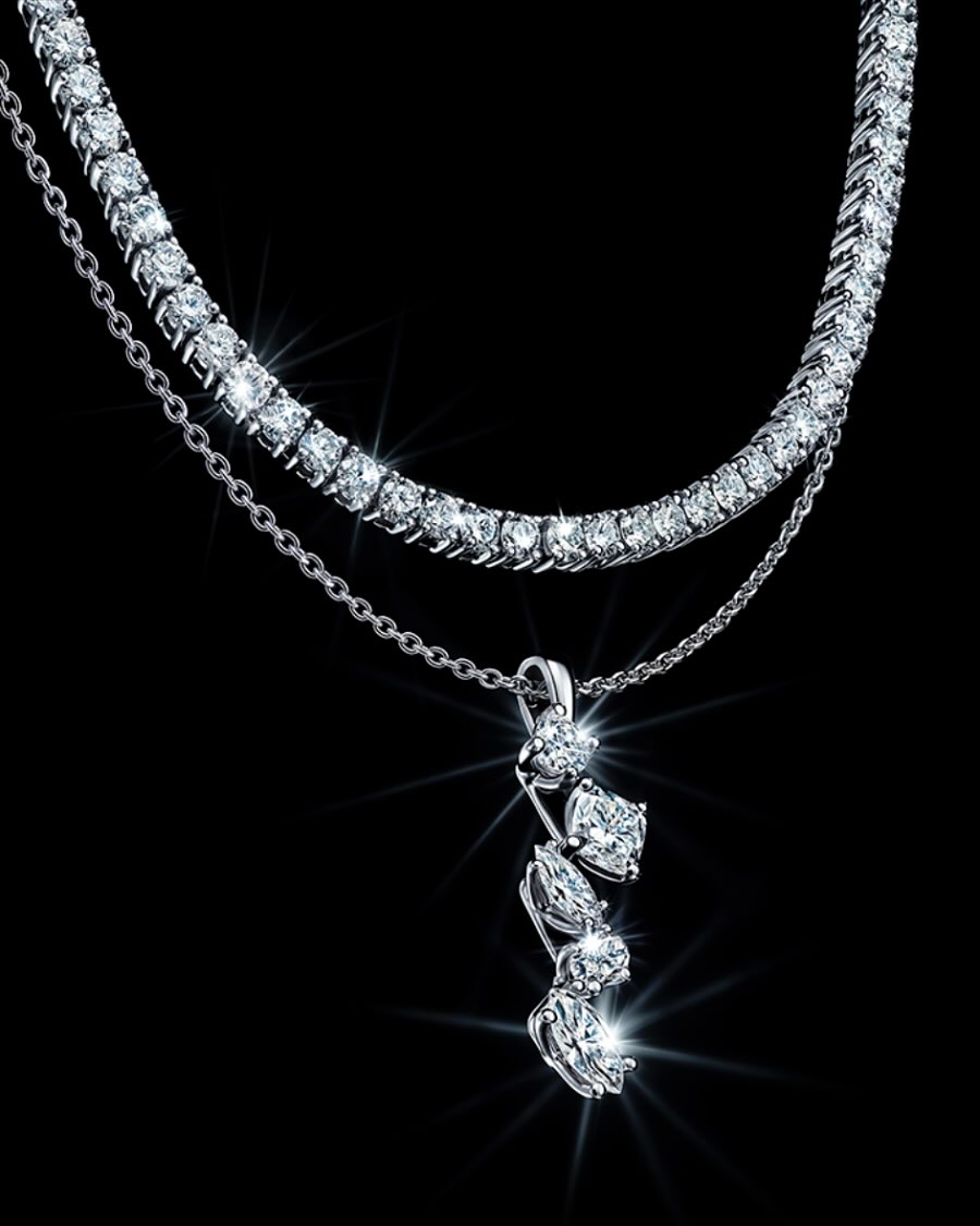 Two-strand diamond necklace on a black backdrop. Features a tennis necklace design and a pendant necklace with cascading stones.