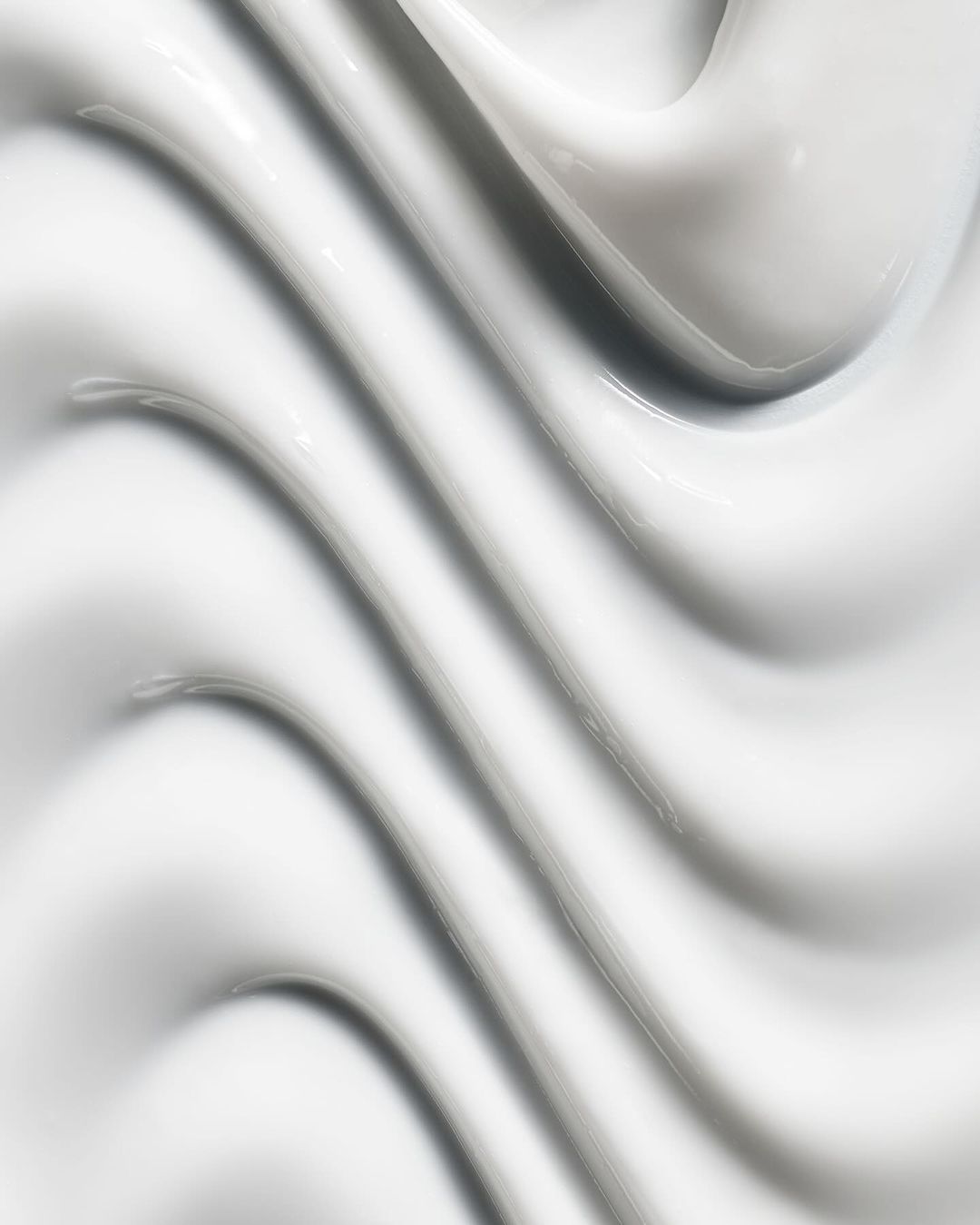 Close-up abstract texture of thick, smooth, glossy white cream or lotion with curving ridges and shadows filling the entire frame.