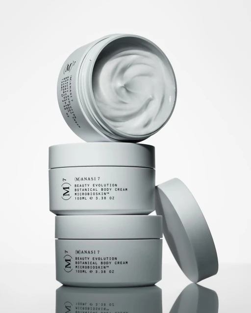 Stacked containers of Manasi 7 botanical body cream with visible product. The stack is positioned on a white background in studio lighting.