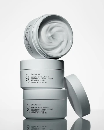 Stacked containers of Manasi 7 botanical body cream with visible product. The stack is positioned on a white background in studio lighting.