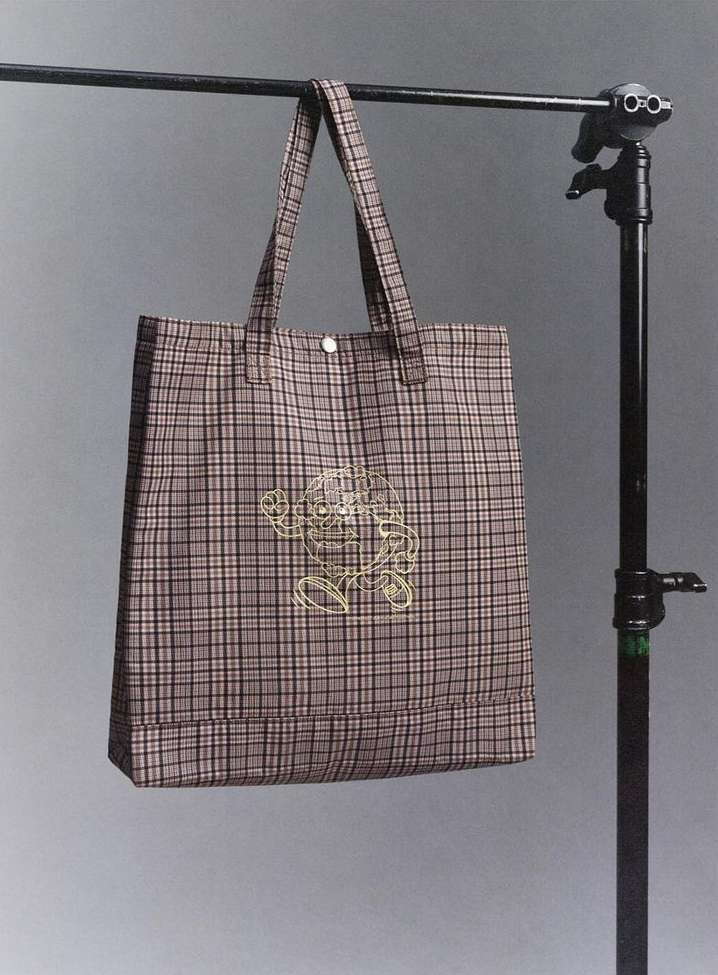 Plaid tote bag with embroidered design hangs from a metal bar with a light gray gradient background, creating a product studio shot.