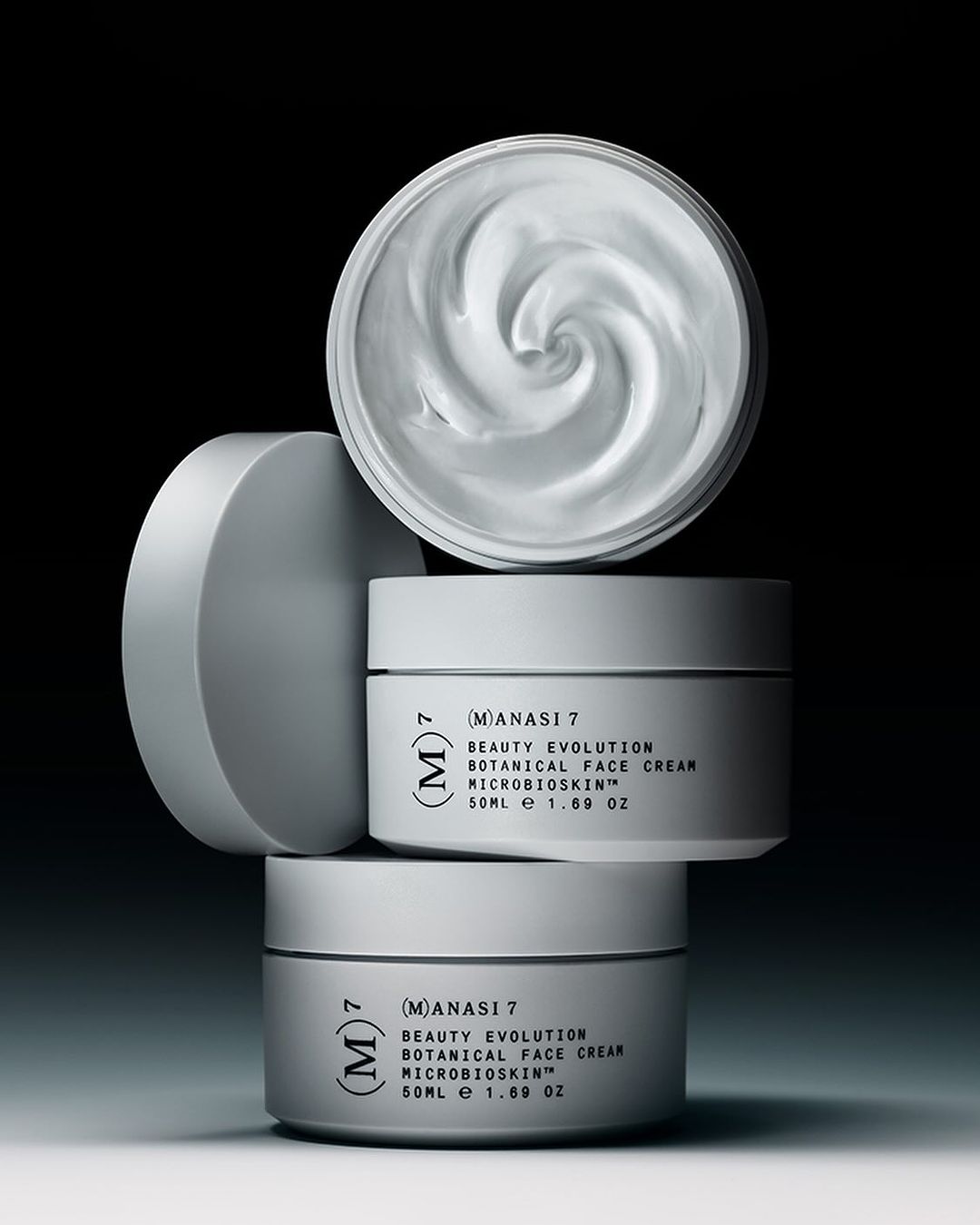 Three white cosmetic jars are stacked. The top jar's lid is off, revealing the creamy contents. A sleek, minimalist product photography.