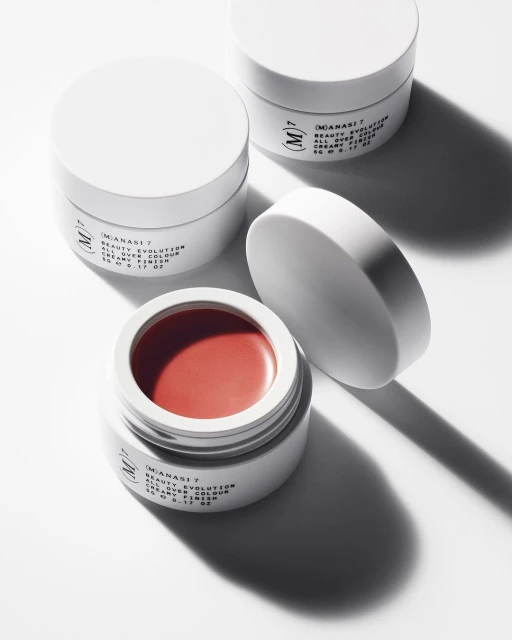 Three white jars of (M)ANASI 7 Beauty Evolution makeup in a minimalist product shot with one jar open showing a coral color.