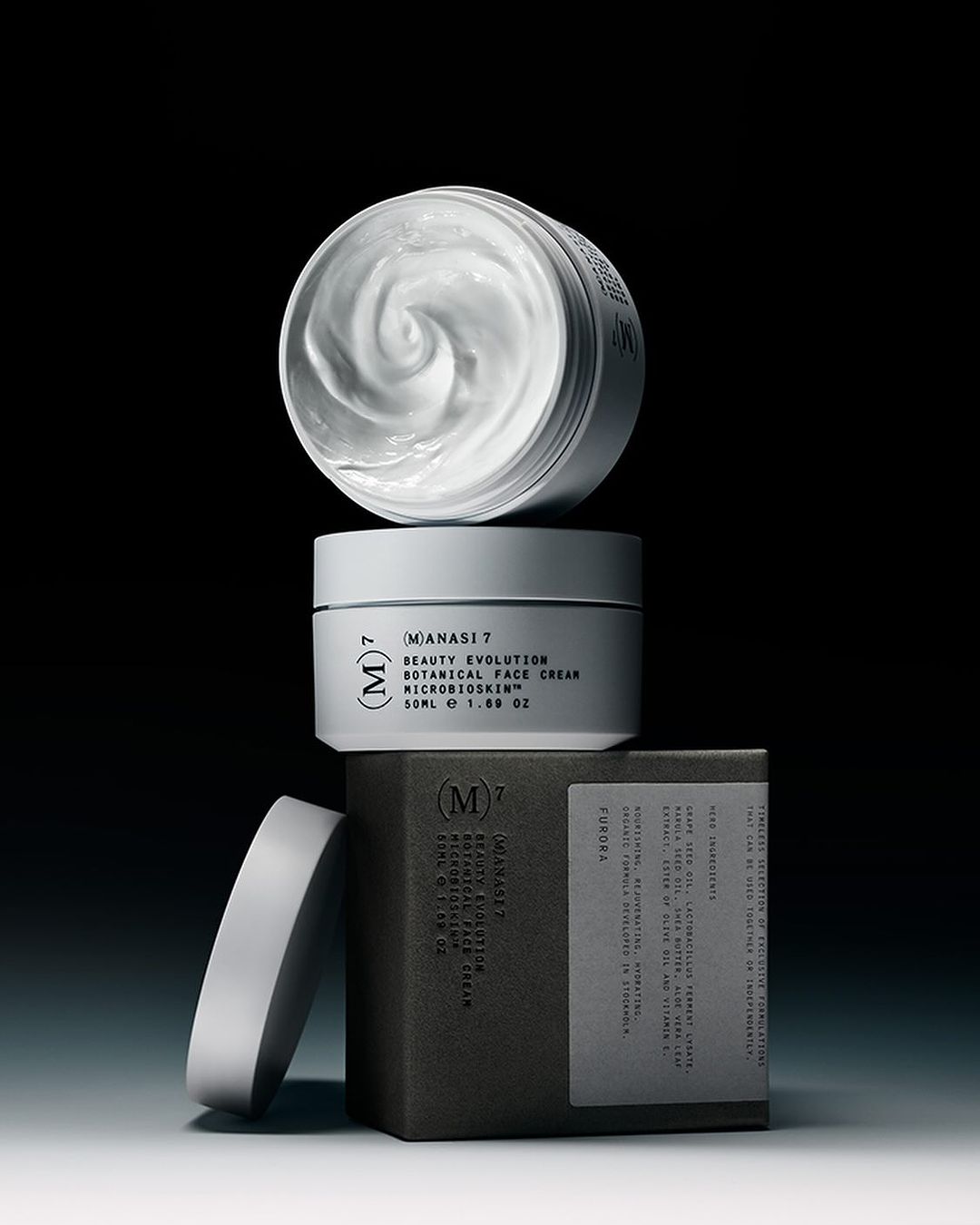 Close-up studio shot of stacked skincare product containers in neutral tones, including cream in open jar and labeled boxes against a black background.
