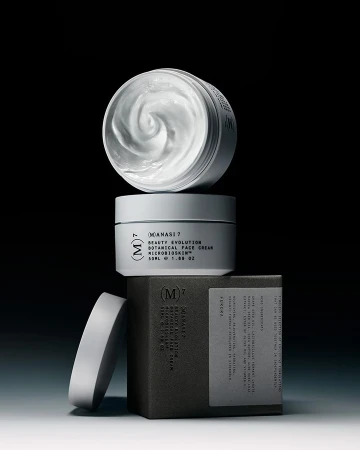 Close-up studio shot of stacked skincare product containers in neutral tones, including cream in open jar and labeled boxes against a black background.