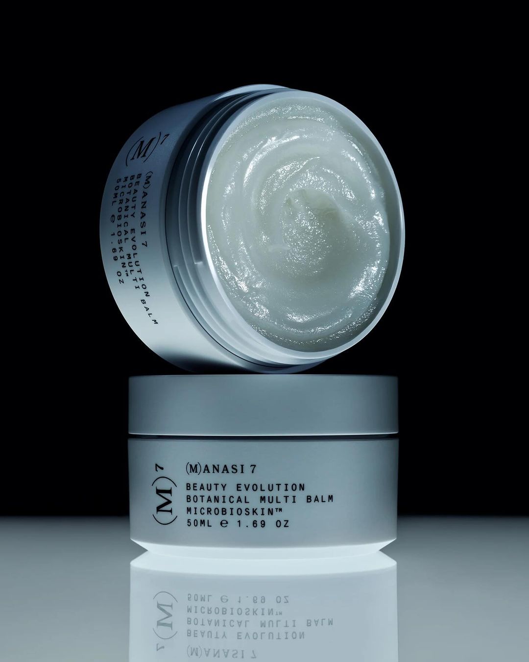 Two stacked jars of Manasi 7 Beauty Evolution Botanical Multi Balm sit on a reflective surface against a dark background. Beauty product shot.