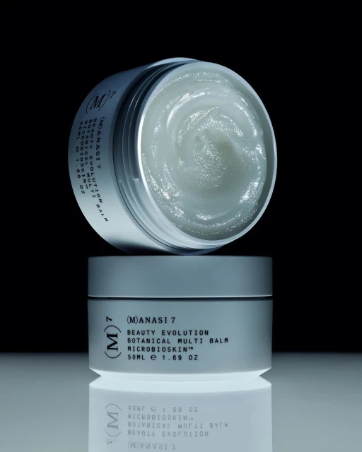 Two stacked jars of Manasi 7 Beauty Evolution Botanical Multi Balm sit on a reflective surface against a dark background. Beauty product shot.