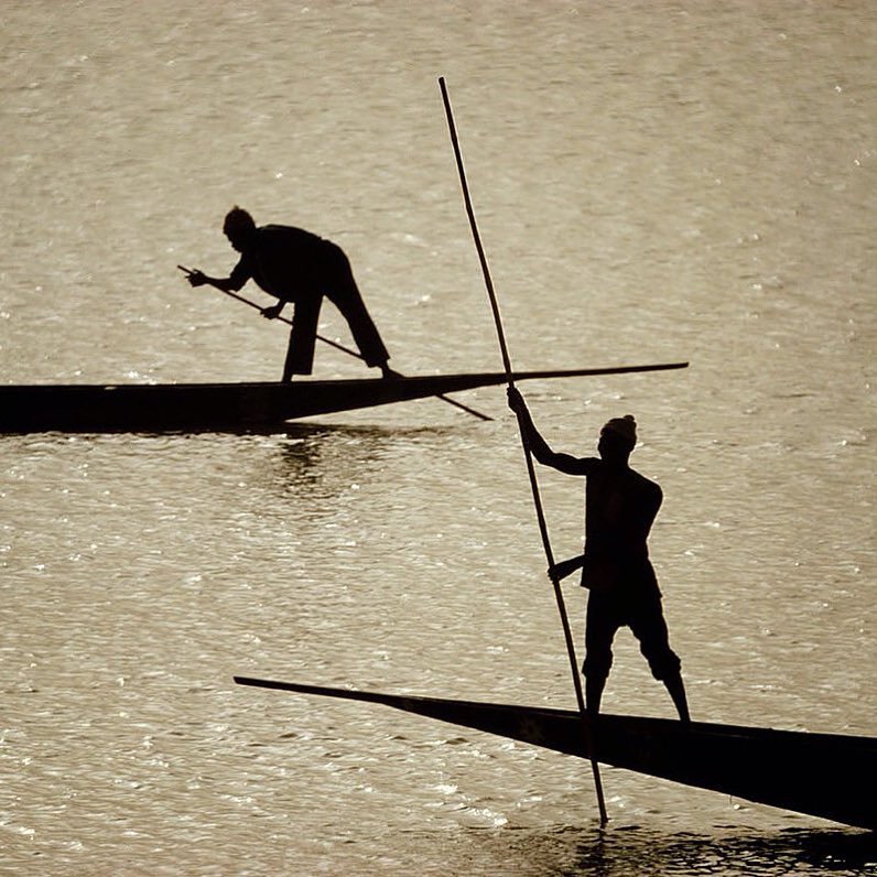 Sepia toned photo of two silhouetted men standing on the ends of long, narrow boats, using poles to navigate through shimmering water.