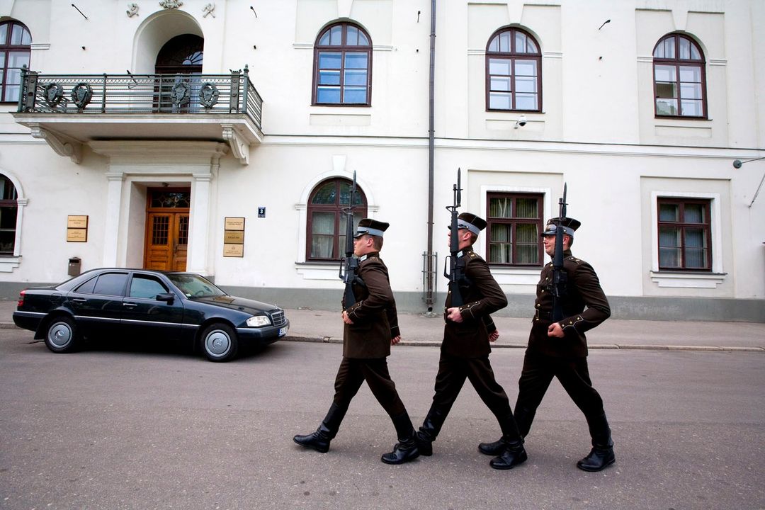 Three guards in uniform march in front of a white building, accompanied by a black car, creating a striking display of formality.