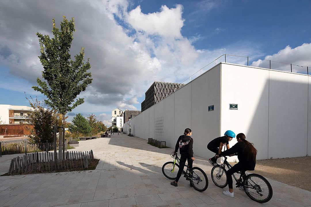 Three people ride bikes on a paved path in an urban park. Landscaping and modern architecture surround the cyclists under a cloudy sky.