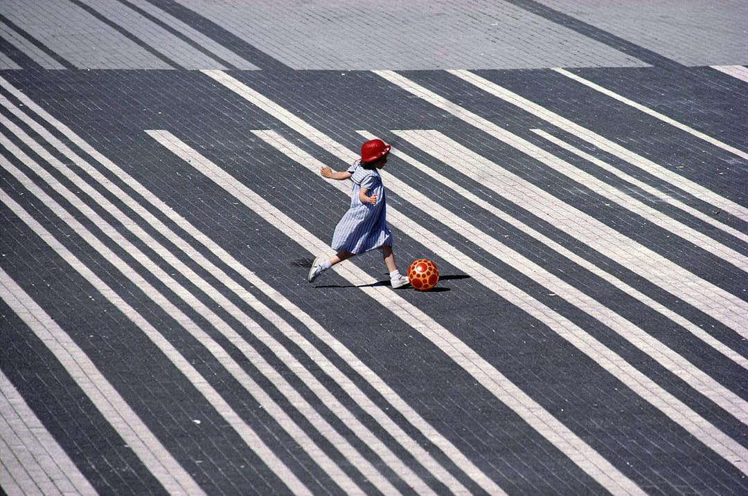 Child in red hat and blue dress kicks patterned ball across striped pavement in sunny outdoor scene. Copy space fills the background.