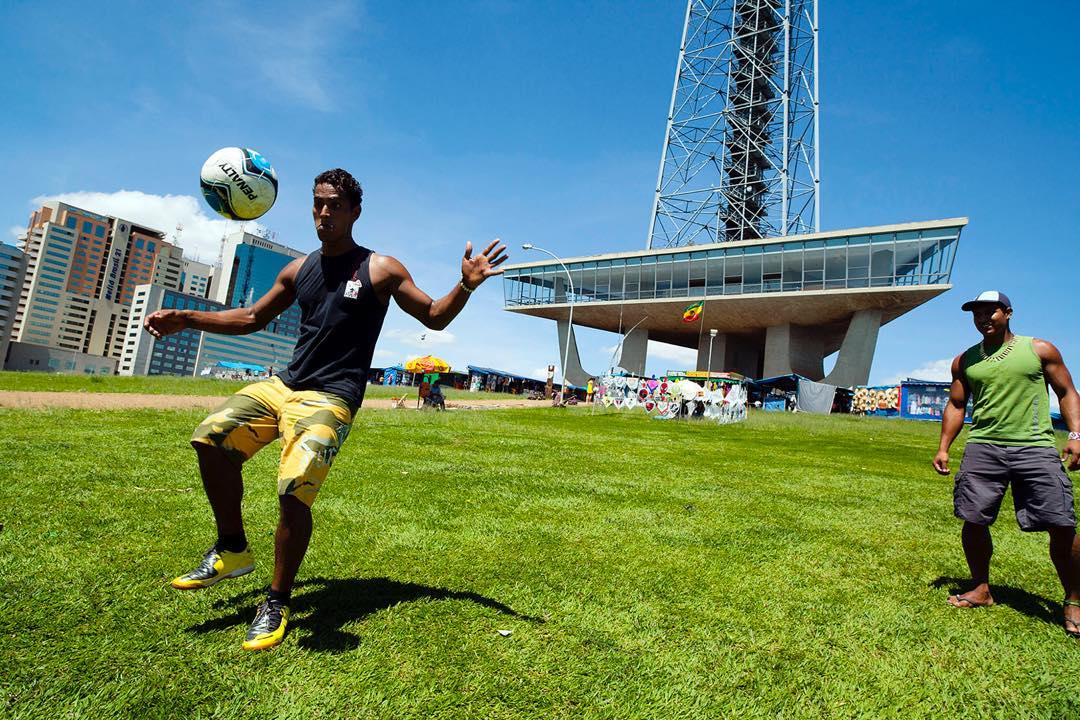 Young man in black tank top balances a soccer ball on his head in an urban park on a sunny day, with a tower structure in the background.