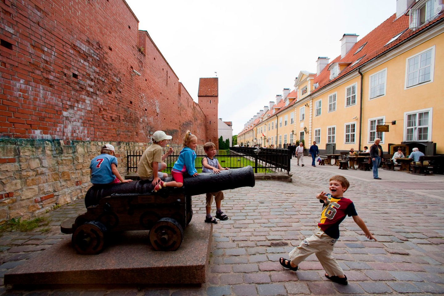 Four children sitting on a cannon in a town square with a young boy excitedly running toward the camera and old buildings lining the street.