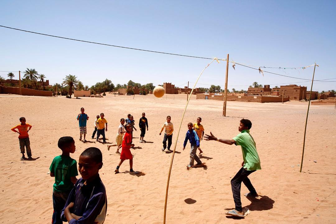 Group of children playing volleyball in a sandy outdoor court with mud brick buildings in the background under a clear blue sky.