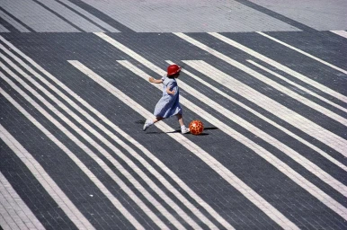 A young girl in a blue dress and red hat kicking an orange patterned ball on a striped stone paved public square.