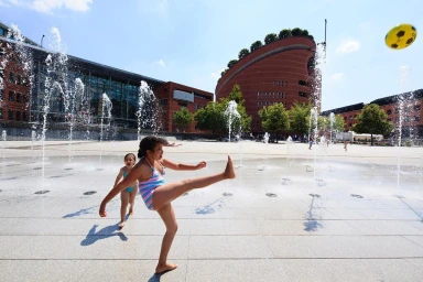 Two young girls playing in a public square with fountains while a yellow ball flies through the air above them.