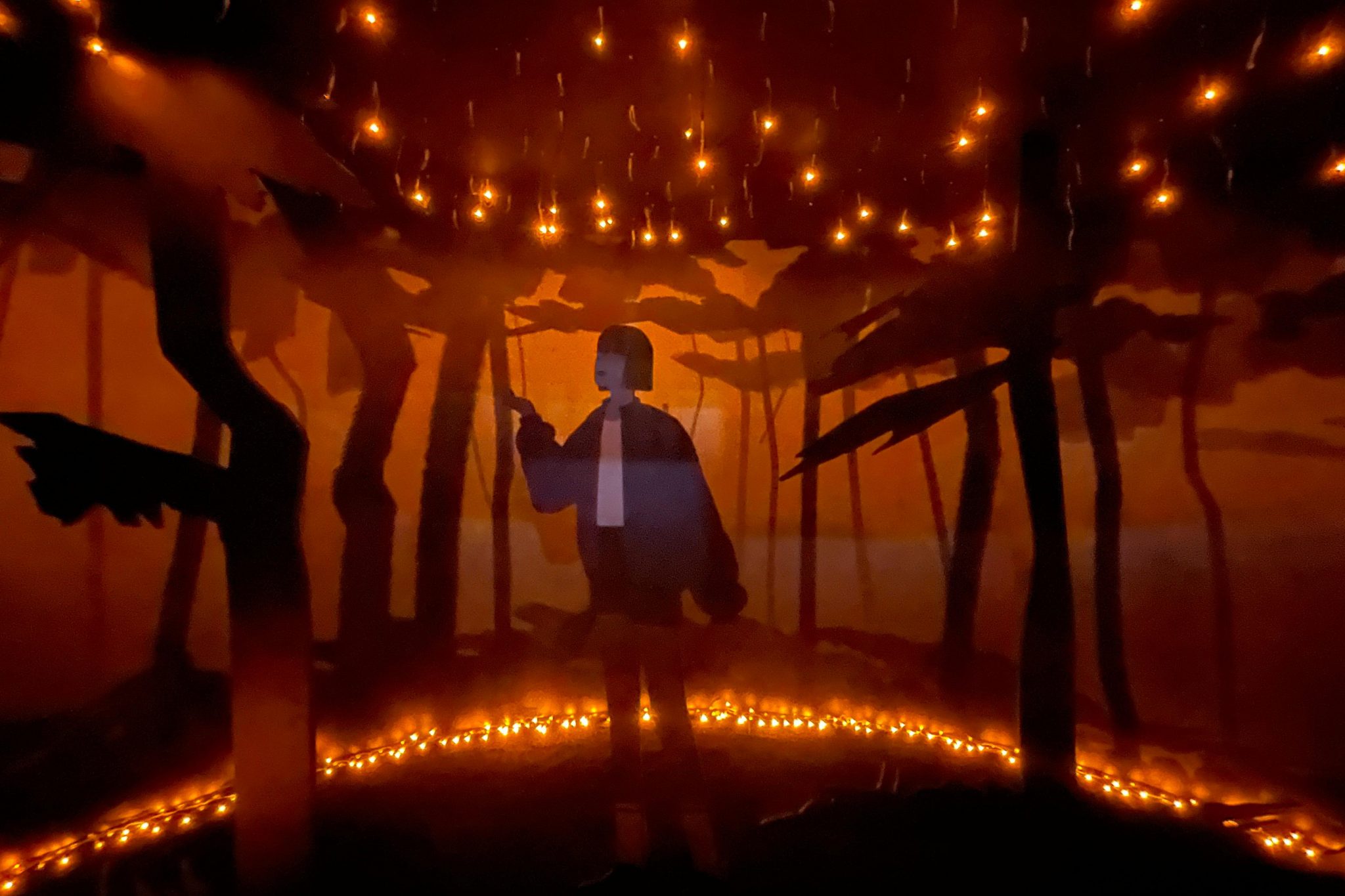 Person stands amid silhouetted trees under string lights, creating a warm, artistic atmosphere in an indoor setting with a forest backdrop.