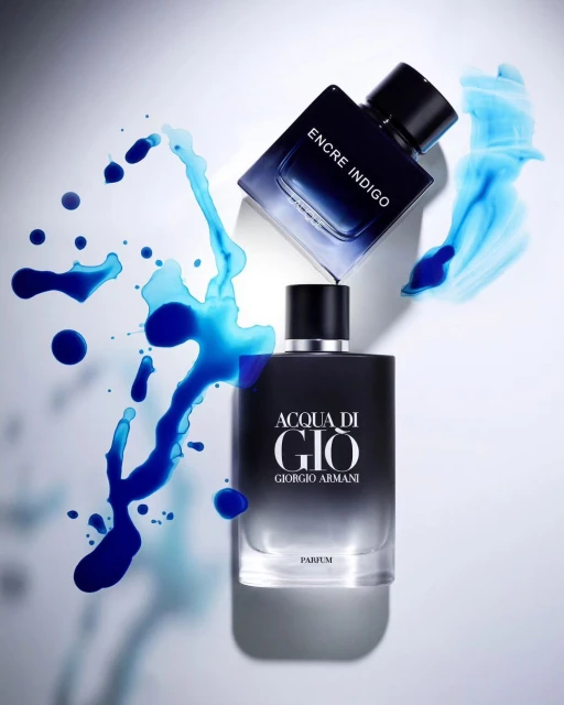 Two perfume bottles, one square and one rectangular, are arranged on a white surface with a splash of blue liquid.