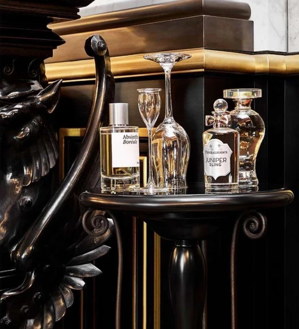 Still life on a black table with Penhaligon's Juniper Sling and Absinthia Borialis perfume bottles, along with clear wine and champagne glasses.