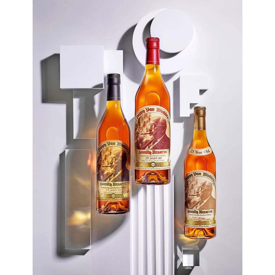 Three bottles of aged bourbon whiskey, each with different colored caps, stand in front of a geometric white background.