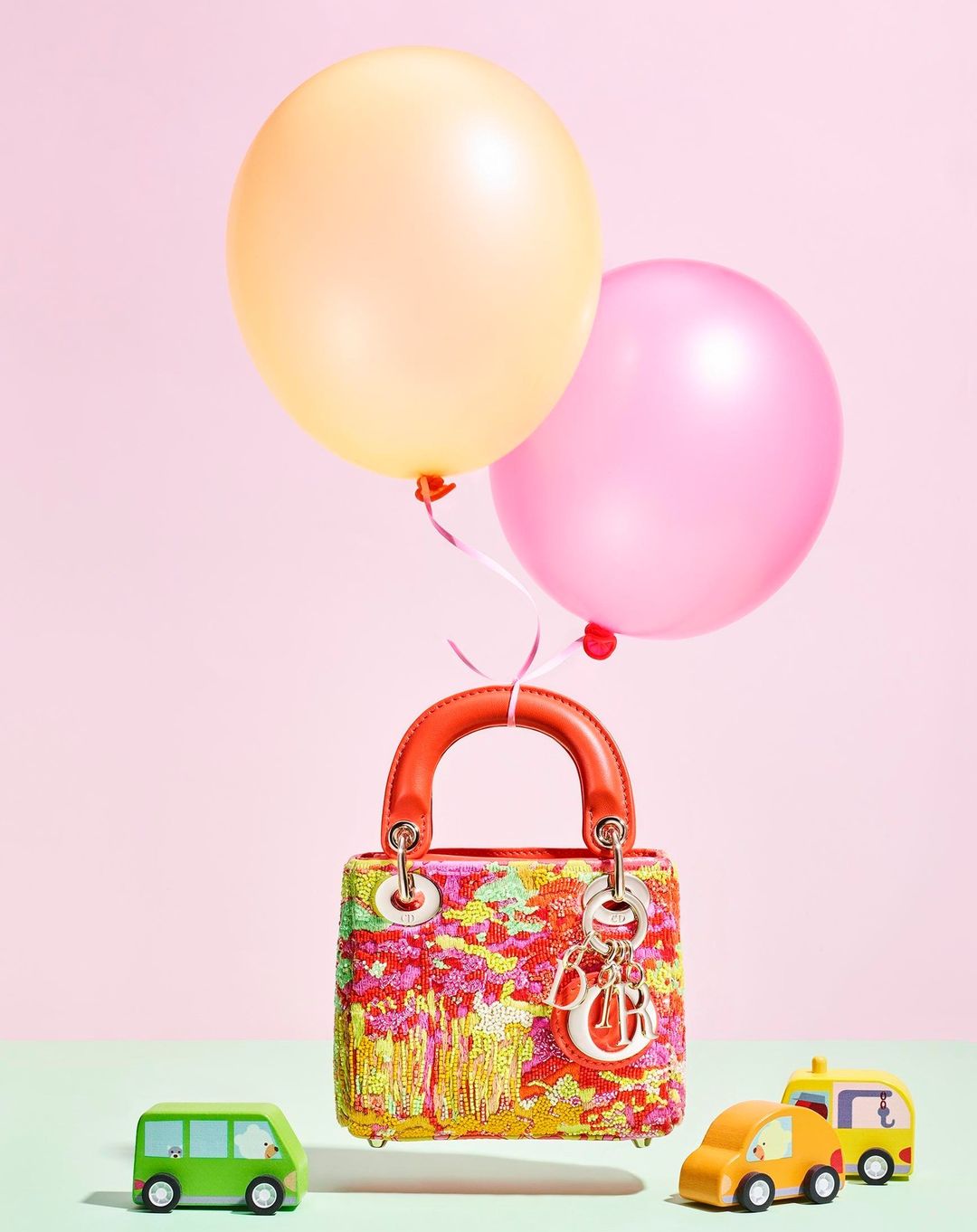 Colorful purse with a red handle is suspended by strings attached to a yellow and pink balloon. Toy cars sit below on a light green surface.