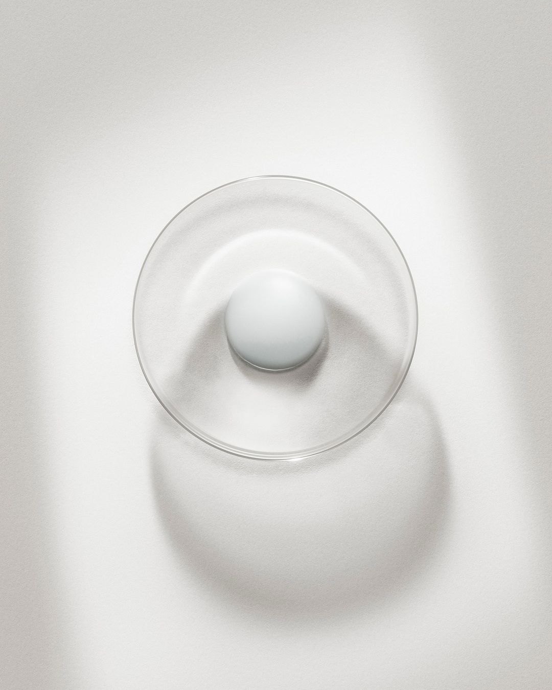 Minimalist interior design featuring a clear glass bowl with a white sphere, set against a clean white backdrop with soft shadows.