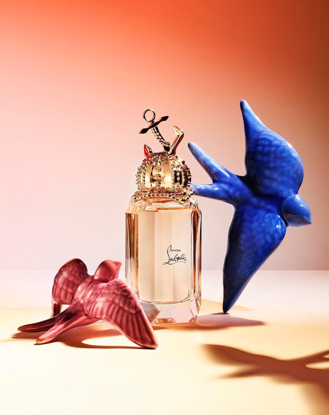 A bottle of Christian Louboutin perfume sits between two ceramic birds, one pink, one blue, against an ombre coral backdrop.