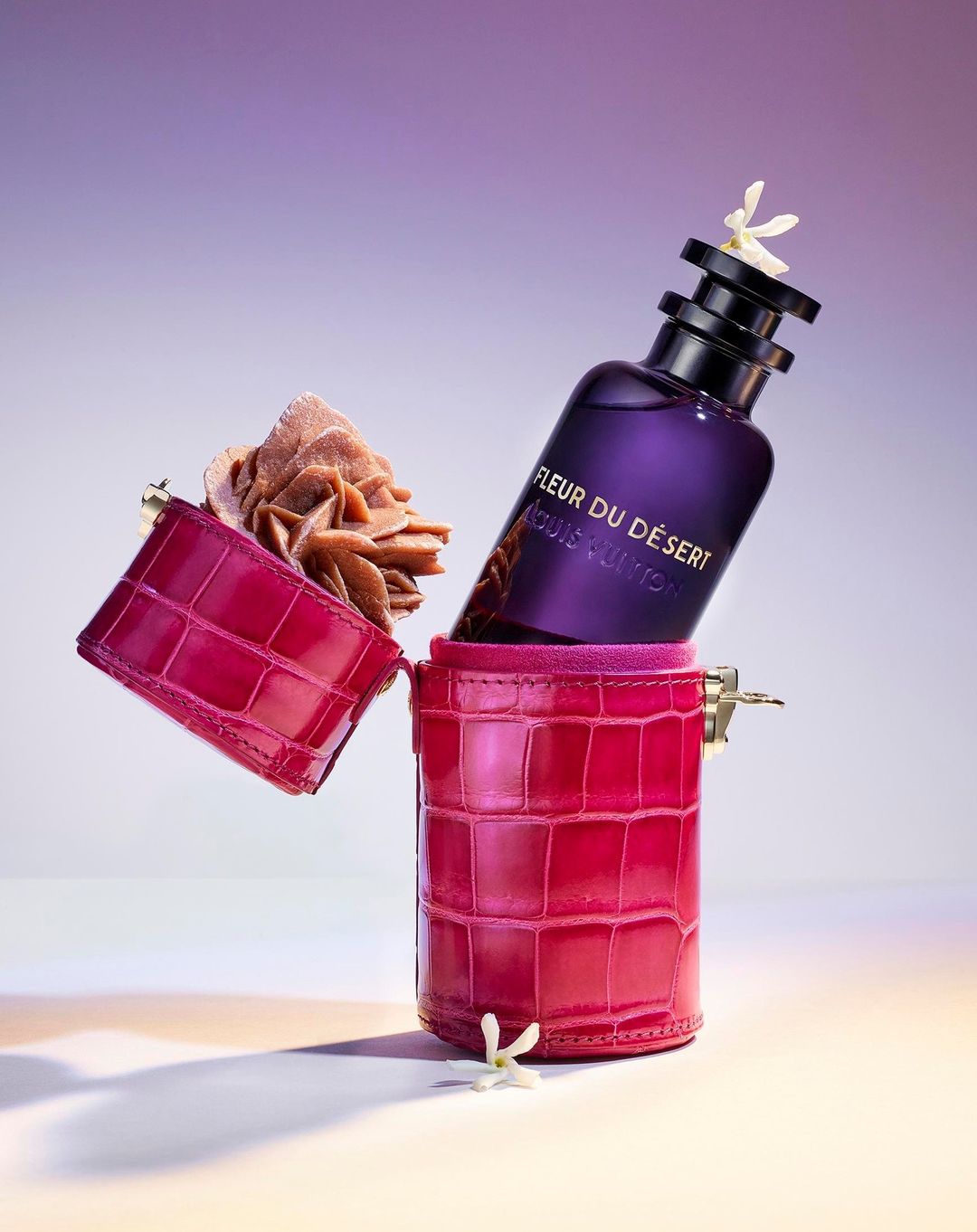 Purple perfume bottle by Louis Vuitton in a pink crocodile case with white flowers, against a gradient light purple background.