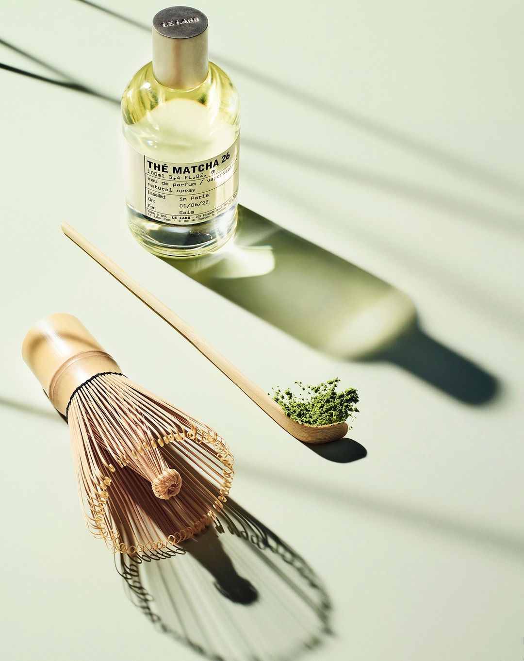 Elevated still life showcasing Le Labo Thé Matcha 26 perfume bottle, matcha powder on a spoon, and a bamboo matcha whisk on a light green backdrop.