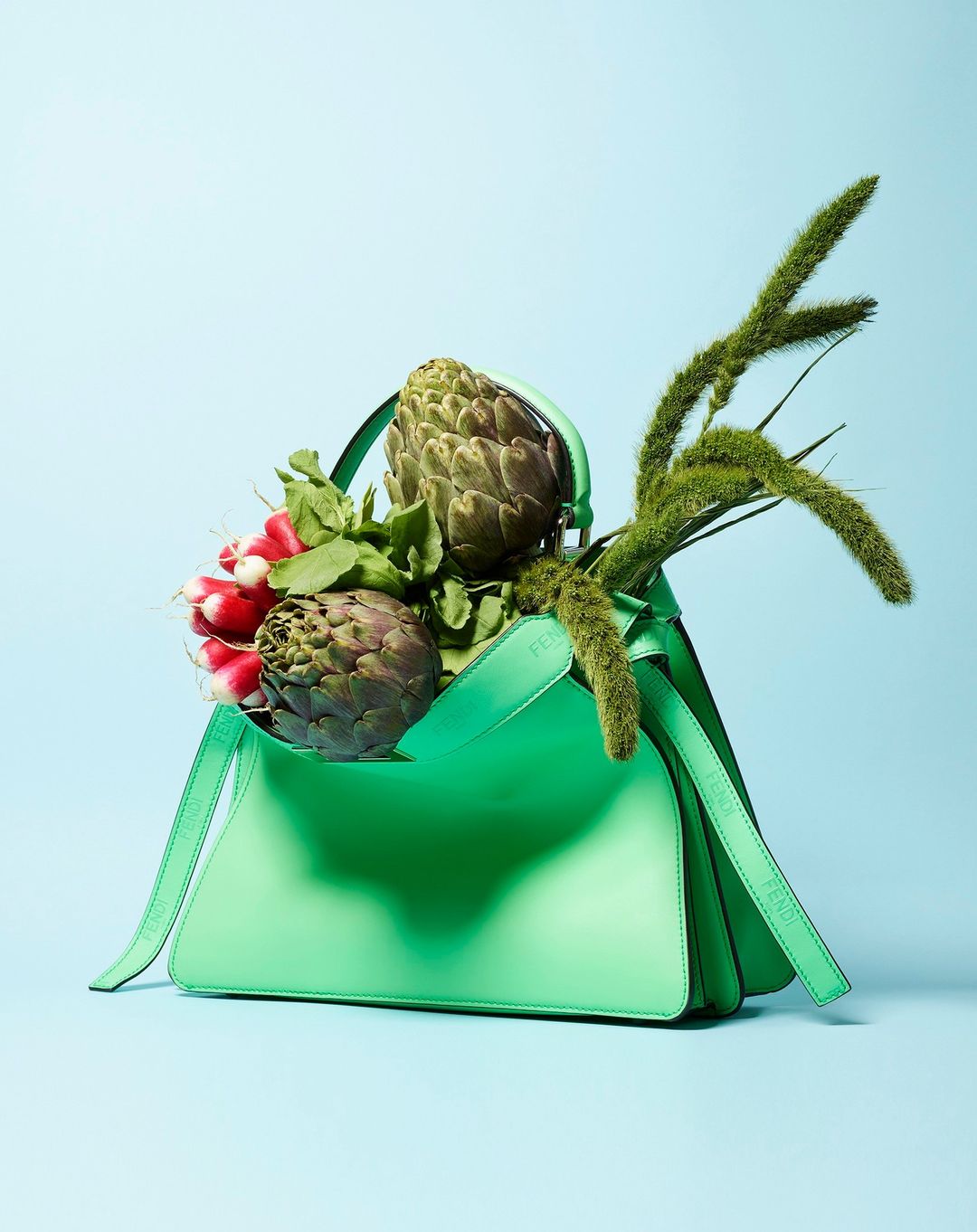 Mint-green leather handbag filled with artichokes, radishes, and green foxtail grass against a light blue background in a studio shot.