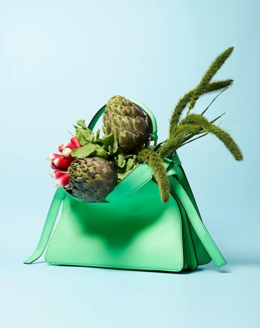 Mint-green leather handbag filled with artichokes, radishes, and green foxtail grass against a light blue background in a studio shot.