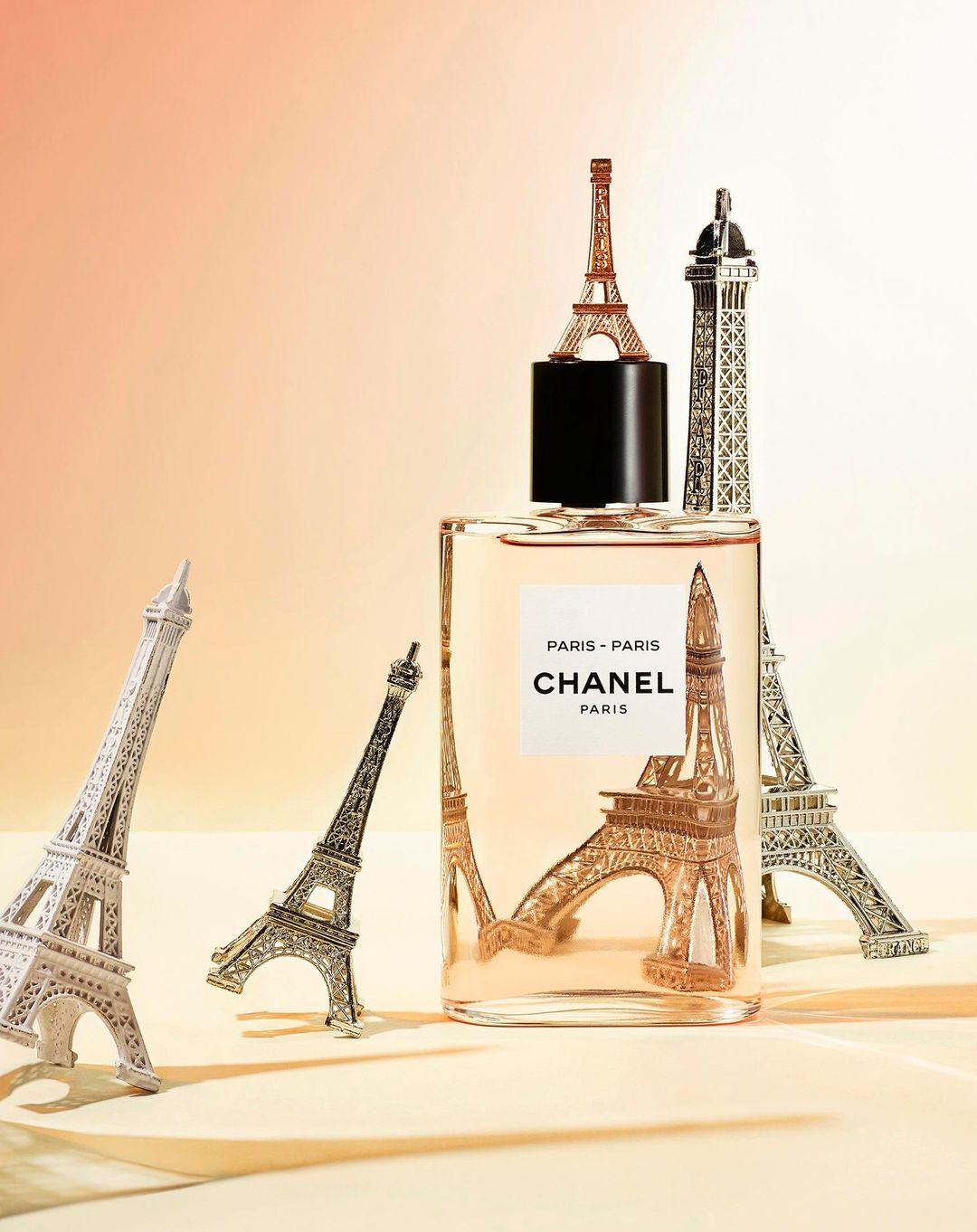 Chanel Paris perfume bottle displayed with Eiffel Tower miniatures on a cream-colored surface against a peach and cream background.