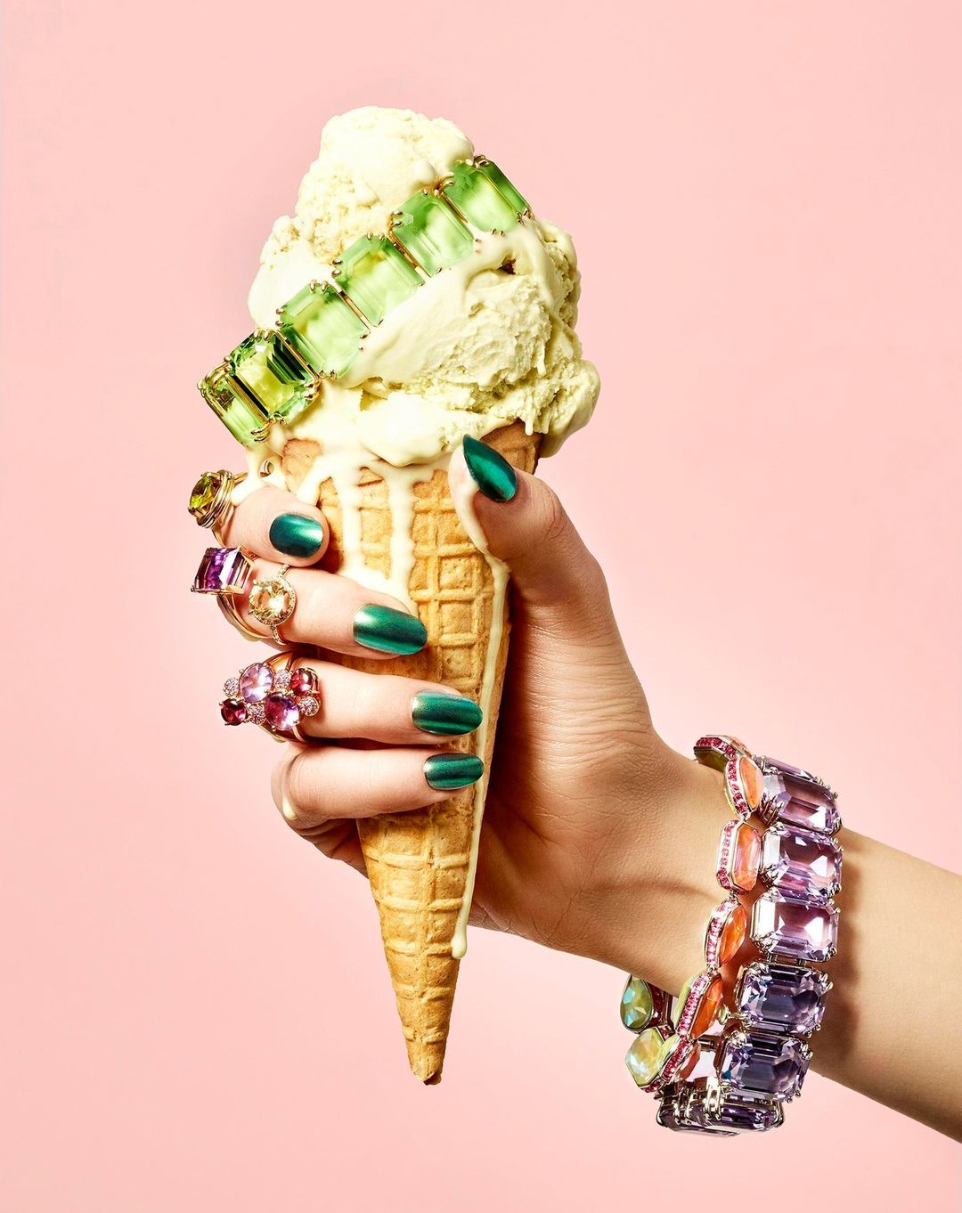 Woman's hand holding an ice cream cone decorated with jewelry against a pink backdrop in a beauty, fashion, and lifestyle setting.