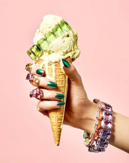 Woman's hand holding an ice cream cone decorated with jewelry against a pink backdrop in a beauty, fashion, and lifestyle setting.
