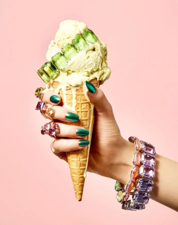 Woman's hand holding an ice cream cone decorated with jewelry against a pink backdrop in a beauty, fashion, and lifestyle setting.