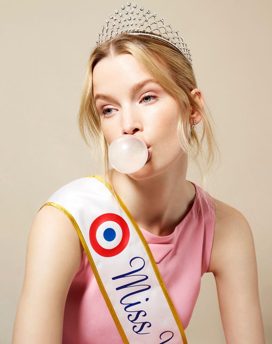 Blonde woman in a pink top blows a bubble with gum while wearing a tiara and a 'Miss' sash in front of a neutral backdrop.