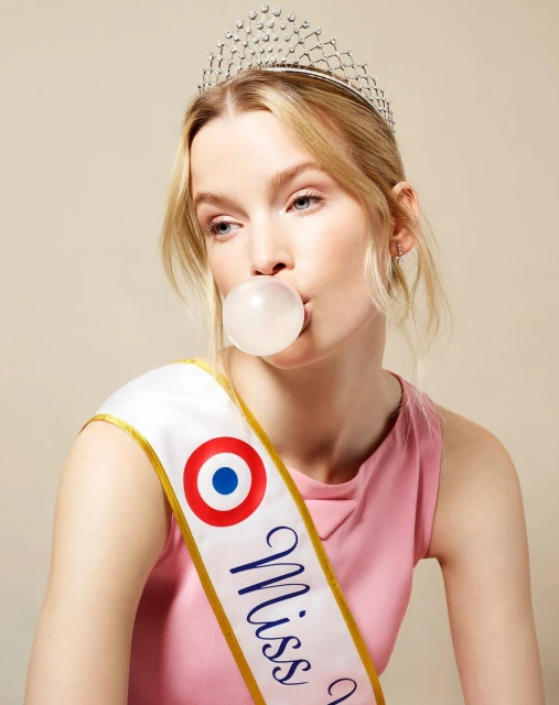 Blonde woman in a pink top blows a bubble with gum while wearing a tiara and a 'Miss' sash in front of a neutral backdrop.