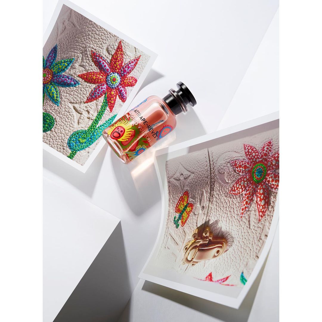 Still life studio shot of a perfume bottle with a black cap, standing among printed images of designer bags with bright floral accents.