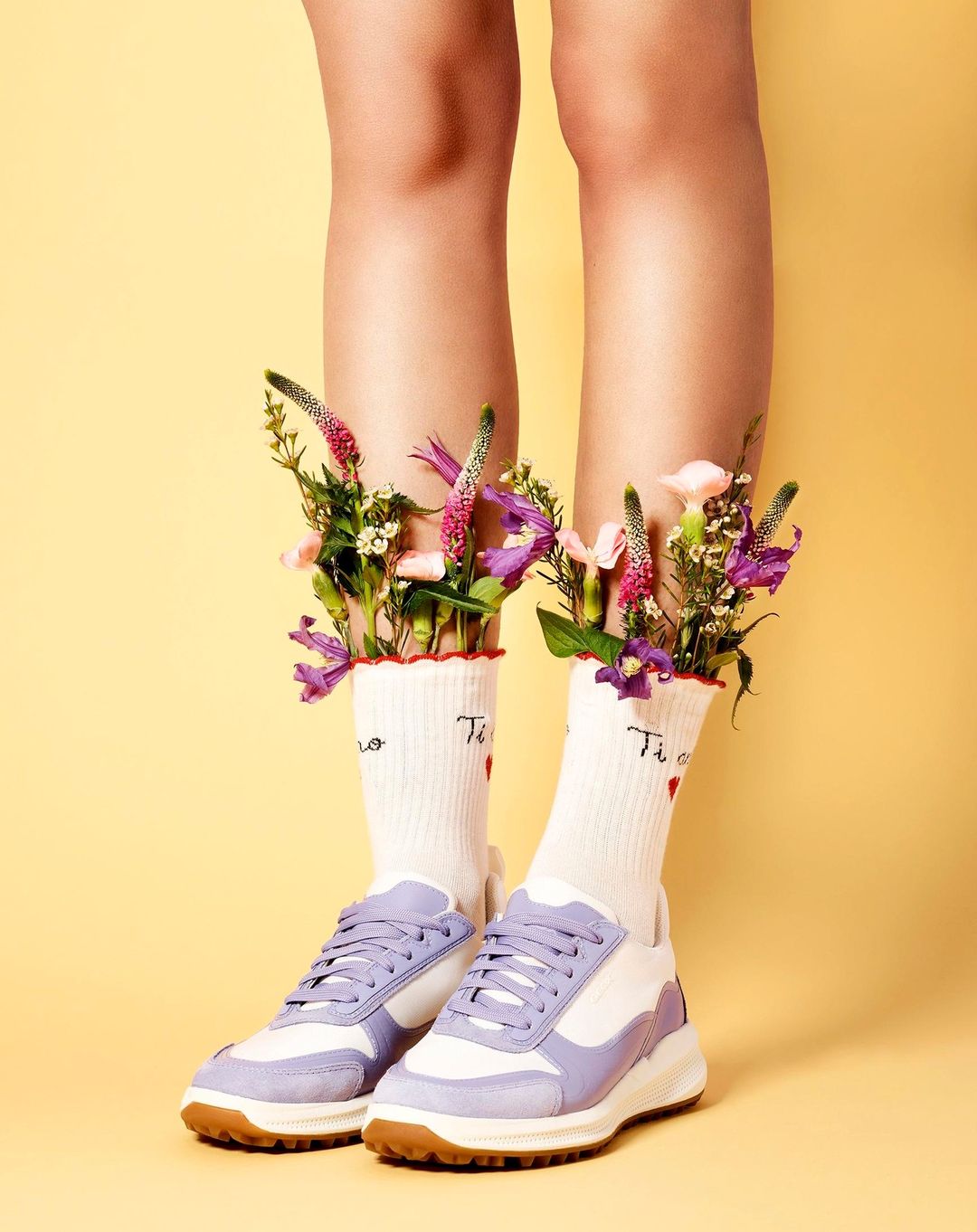 Close-up of bare legs wearing white socks filled with flowers and pastel purple sneakers against a yellow background