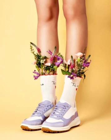 Close-up of bare legs wearing white socks filled with flowers and pastel purple sneakers against a yellow background