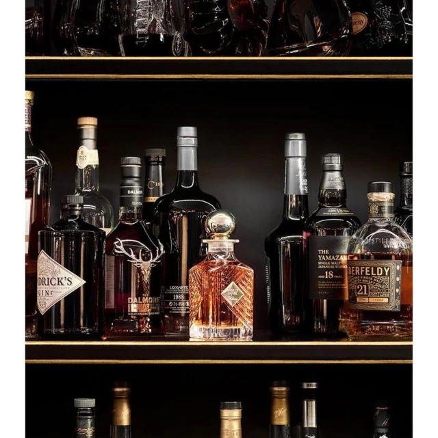 Liquor bottles are displayed on dark shelves with gold trim, featuring various brands, shapes, and amber liquid contents.
