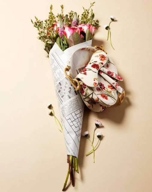Floral purse sits on a bouquet of pink roses wrapped in newspaper, accompanied by small daisies on a light beige background.
