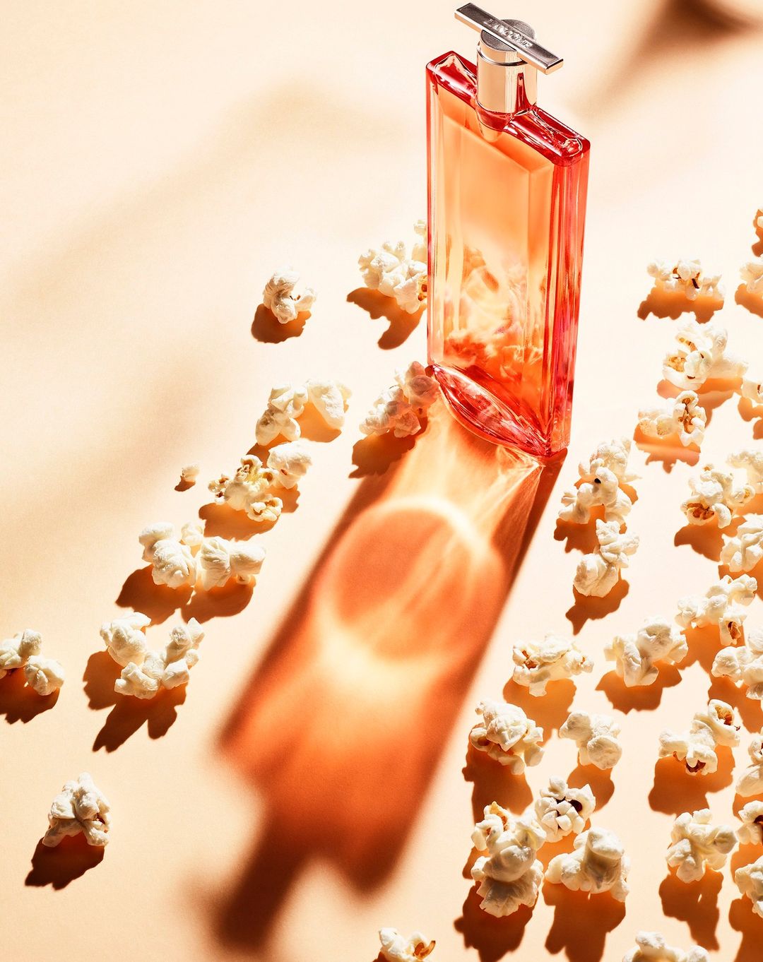 A perfume bottle casts a shadow on a peach-colored surface surrounded by scattered popcorn, creating a beauty still life.