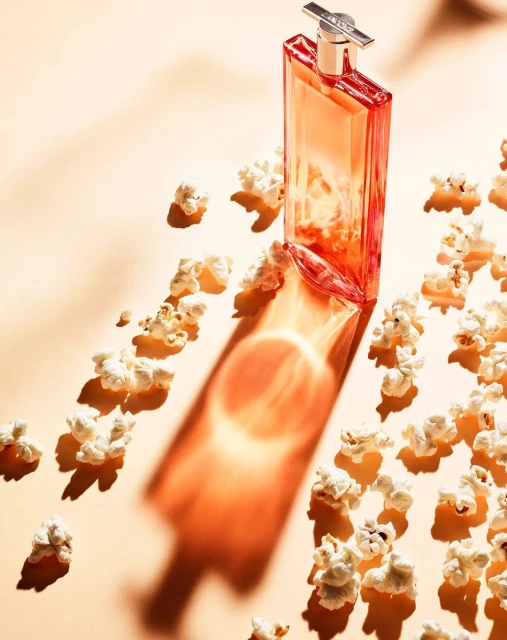 A perfume bottle casts a shadow on a peach-colored surface surrounded by scattered popcorn, creating a beauty still life.