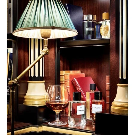 Still life of perfume bottles with books, a cognac glass, and a table lamp in a dimly lit, warmly colored interior setting.