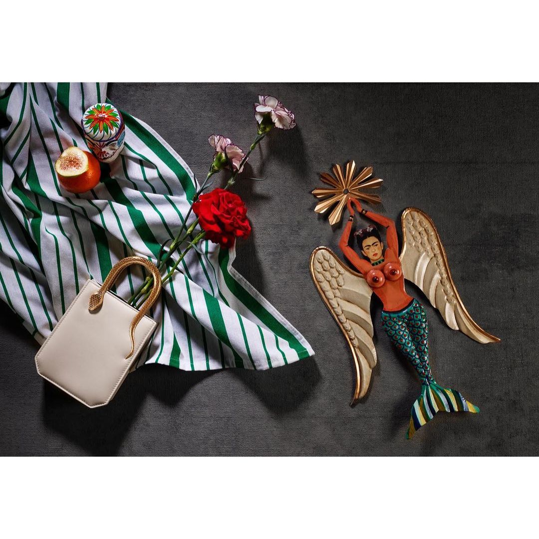 Still life composition features a mermaid figure, handbag, flowers, striped cloth, and fruit on a grey surface, creating a whimsical scene.